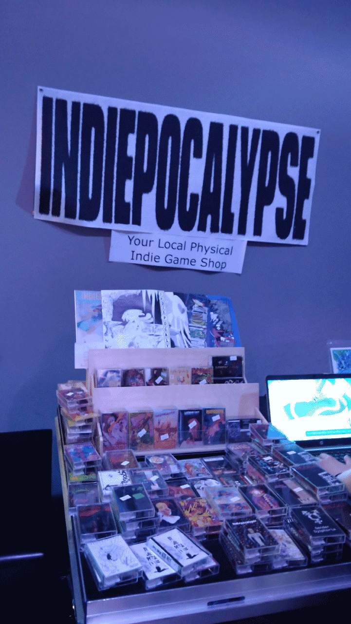 A vertical photo of a table with many cassette tape cases on it, as well as a laptop that a booth visitor is playing a game on. A big horizontal poster with words "INDIEPOCALYPSE" and "Your Local Physical Indie Game Shop" hangs on the wall behind the table.